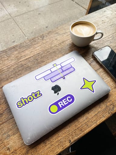 Violet stickers on a laptop lid in a West Village café: the shotz wordmark, an editing timeline with a lime playhead, a REC pill and a lime star