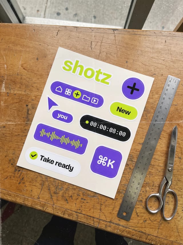 A kiss-cut sheet of shotz stickers on a Chinatown print shop counter beside a steel ruler and scissors: the wordmark, a lime plus button, the tab bar, a pointer reading you, a New badge, a timecode, a waveform, a Take ready toast and a ⌘K key