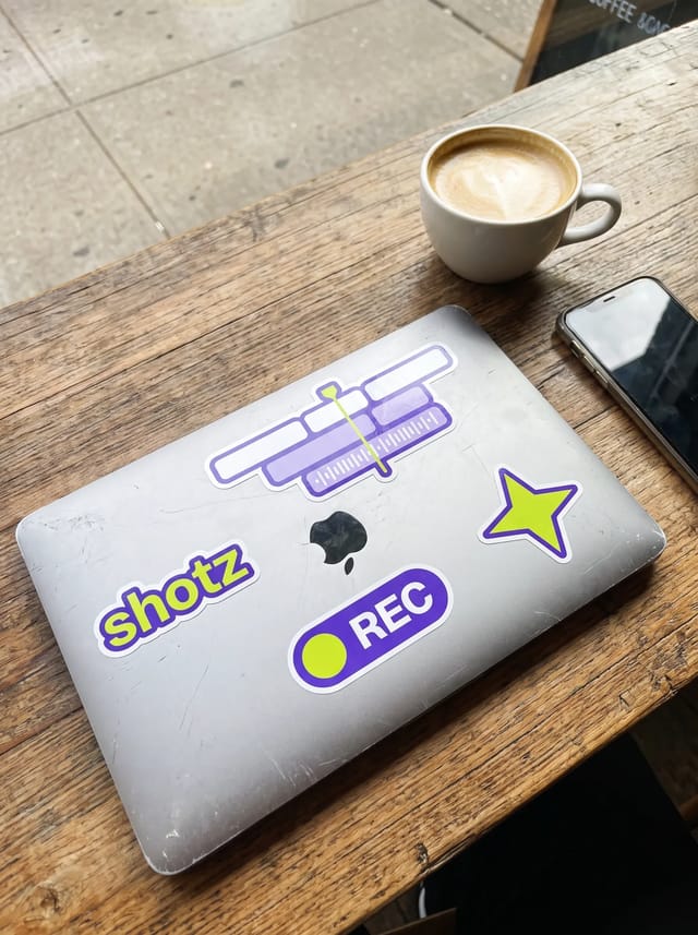 Violet stickers on a laptop lid in a West Village café: the shotz wordmark, an editing timeline with a lime playhead, a REC pill and a lime star