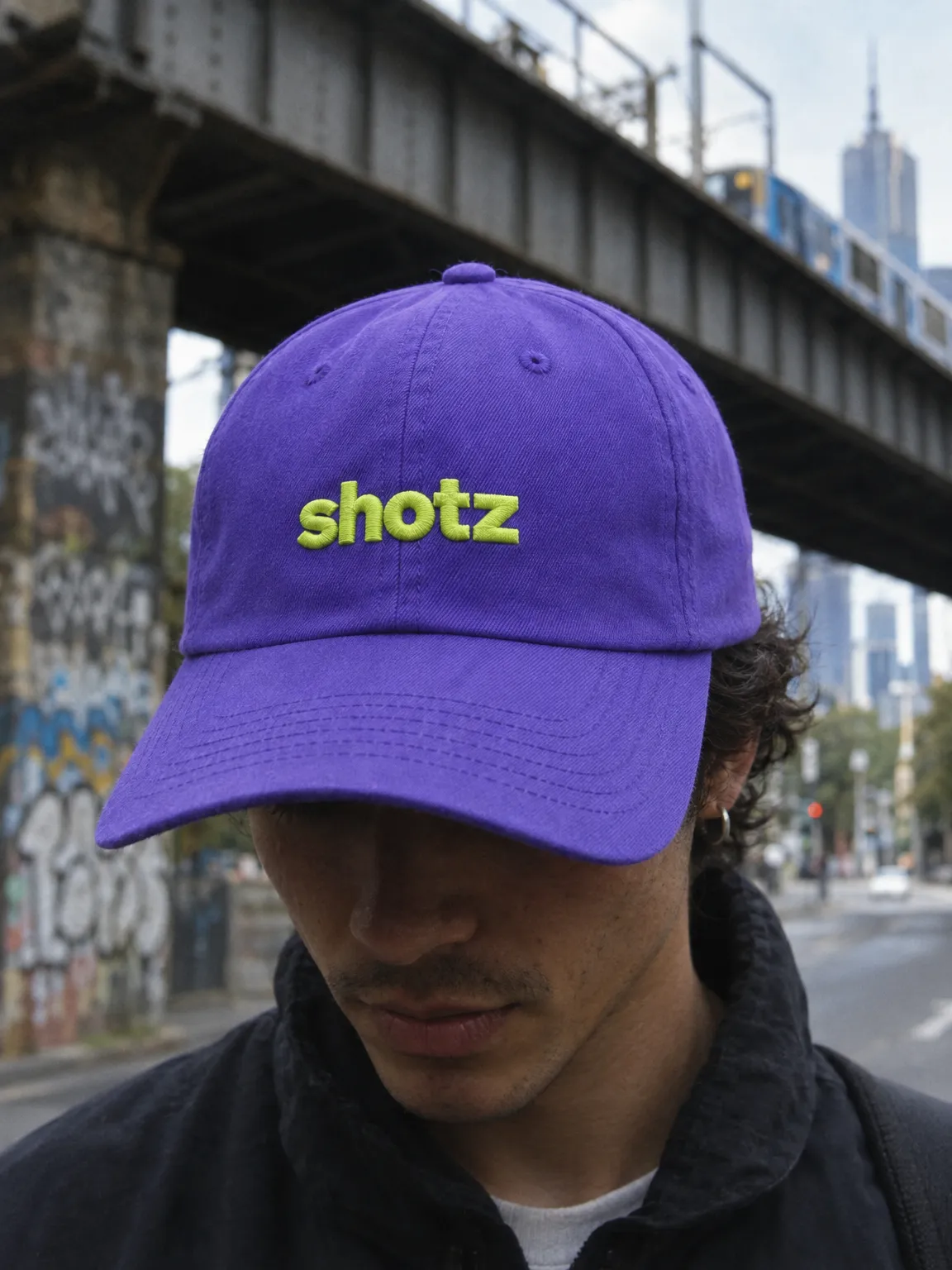 Violet cap with a raised lime shotz embroidery, worn under a train bridge