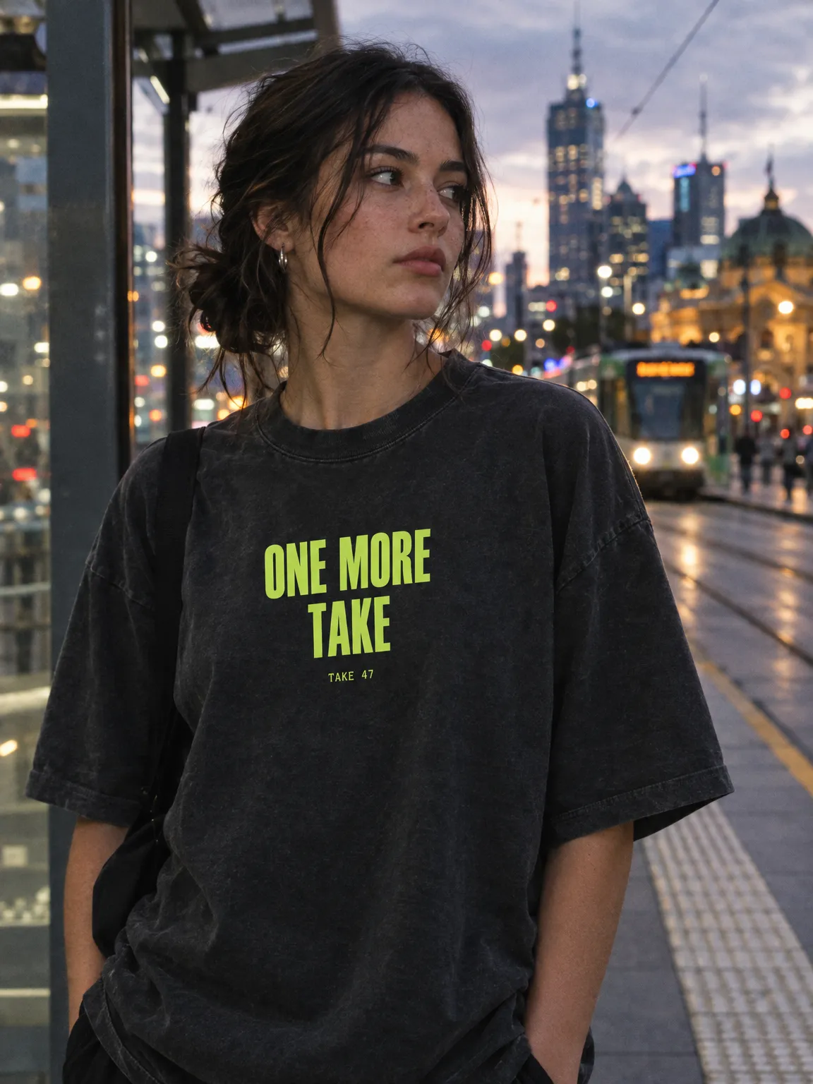 Washed black tee printed ONE MORE TAKE in lime, worn at a tram stop at dusk